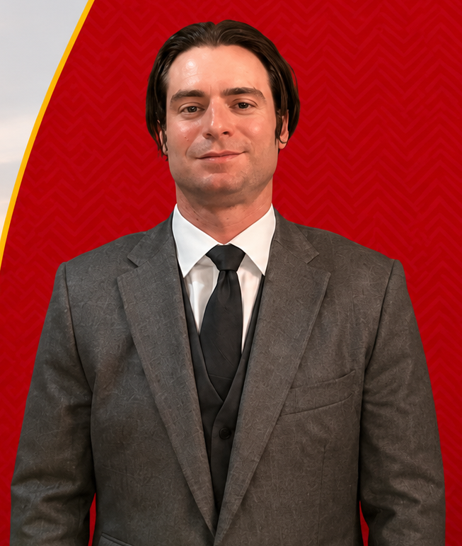 Man in a gray suit and black tie standing against a red patterned background